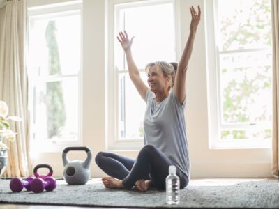 Smiling mature woman with arms raised doing yoga. Fit female is sitting by exercise equipment on rug. She is wearing sports clothing at home.